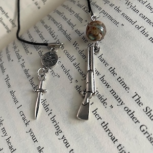 May include: A black cord necklace with three silver charms: a compass, a dagger, and a rifle. The necklace is laying on a book with text about Grisha and the Shura.