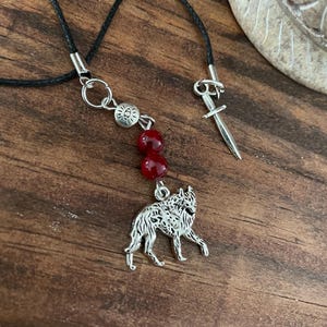 May include: A black cord necklace with a silver wolf pendant, red beads, and a decorative sword clasp. The wolf pendant is detailed and the beads are a deep red color. The necklace is displayed on a wooden surface.