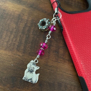 May include: A silver cat charm with a heart, a pink beaded strand, and a small donut charm. The charms are attached to a black cord, next to a red phone case.