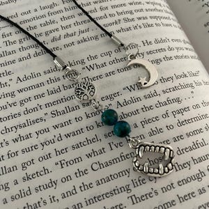 May include: A collection of silver-toned bookmarks with various charms, including a tree of life, a crescent moon with a star, and a set of vampire teeth. The bookmarks are strung on black cords and adorned with teal beads.