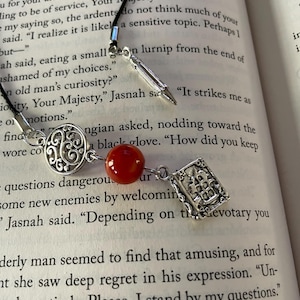 May include: A silver bookmark with a red gemstone charm, a silver quill charm, and a silver book charm. The bookmark is hanging from a black cord. The bookmark is on top of a book with text that reads "It strikes me as" and "How did you keep questions dangerous".