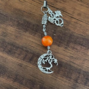 May include: A silver charm with a dragon, a decorative bead, and a crescent moon with a tree design. The charm is attached to a black cord. The bead is orange.