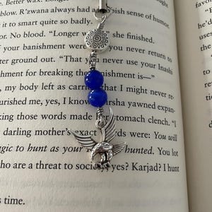 May include: A silver-toned bookmark with a black cord, featuring a decorative sunburst charm, two blue beads, and a detailed eagle pendant. The bookmark is designed to mark pages in a book.