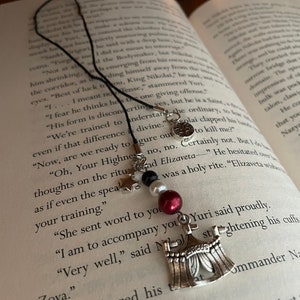 May include: A silver bookmark with a tent design, black beads, a white pearl, and a red bead. The bookmark is hanging from a black string and is resting on a book with text about a saint.