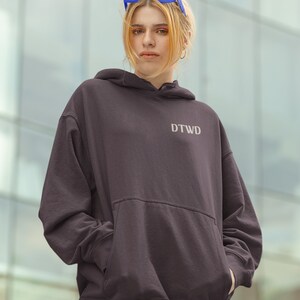 May include: A dark grey hoodie with a large pocket and the text "DTWD" embroidered on the front. The model is wearing the hoodie with blue sunglasses.