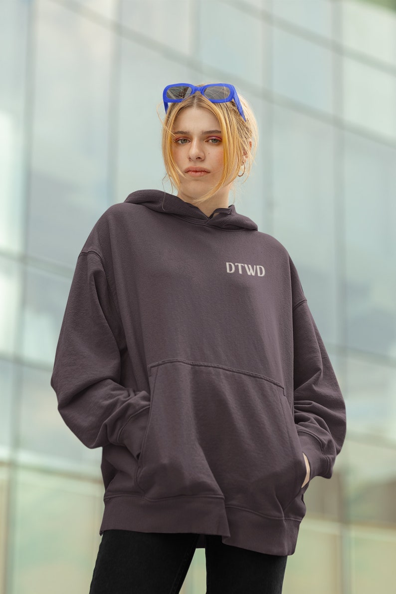 May include: A dark grey hoodie with a large pocket and the text "DTWD" embroidered on the front. The model is wearing the hoodie with blue sunglasses.