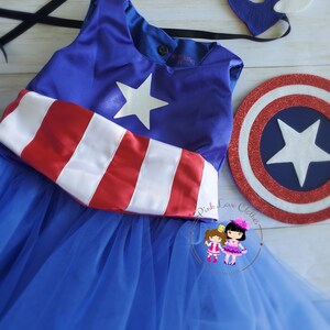 May include: A blue and red superhero costume set, including a dress with a white star and red and white stripes, a matching mask, and a shield. The dress has a blue tulle skirt. The set is designed for children.