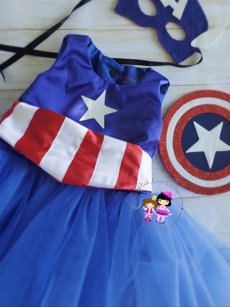 May include: A blue and red superhero costume set, including a dress with a white star and red and white stripes, a matching mask, and a shield. The dress has a blue tulle skirt. The set is designed for children.