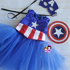 May include: A blue Captain America-themed costume set. The set includes a blue dress with a white star and red and white stripes, a matching mask with the letter "A", and a shield. The dress has a blue tulle skirt.