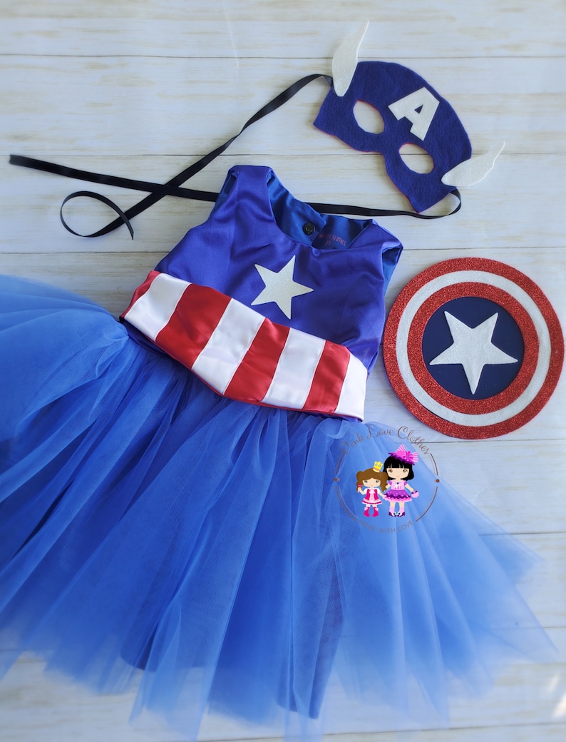 May include: A blue Captain America-themed costume set. The set includes a blue dress with a white star and red and white stripes, a matching mask with the letter "A", and a shield. The dress has a blue tulle skirt.