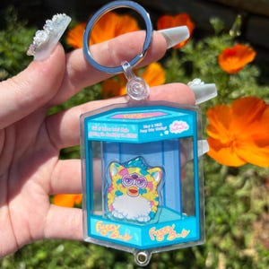 Op de afbeelding: Een heldere acryl sleutelhanger met een kleurrijk Furby-achtig karakter in een blauwe en heldere behuizing. De sleutelhanger heeft een zilveren metalen ring. De tekst "Fuzzy Baby" staat op de behuizing.