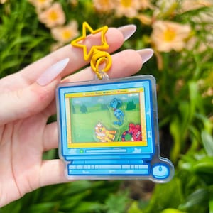 May include: A clear acrylic keychain shaped like a vintage computer monitor with a yellow star-shaped clasp. The screen features a green landscape graphic with three small, colorful, floating character charms inside the transparent casing.