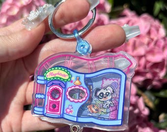 Puede incluir: Un llavero acrílico transparente con un diseño de casa de juegos en miniatura. El llavero tiene un anillo plateado y un conector de plástico azul. La casa de juegos tiene una puerta rosa, un marco azul y un perro y un pez de dibujos animados en el interior.