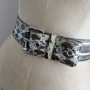 Puede incluir: Un cinturón ancho de piel de serpiente blanco y negro con hebilla plateada y ojales. El cinturón se lleva en un maniquí.