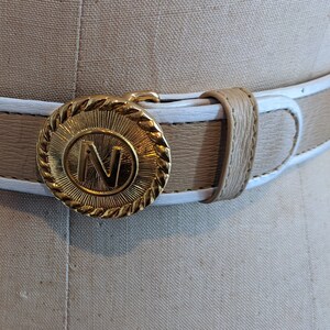 May include: A tan leather belt with a white edge and a gold buckle featuring a raised letter "N" in the center.