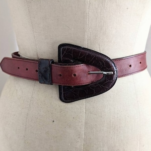 May include: A burgundy leather belt with a large, dark brown, textured buckle. The buckle has a silver metal pin.