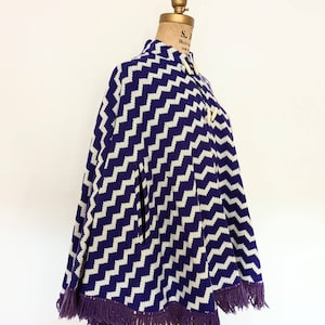 May include: Purple and white chevron patterned knitted cape with a purple fringe trim. The cape has a high collar and two buttons.