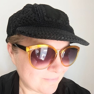 Könnte beinhalten: Eine Person trägt eine schwarze Häkelmütze mit breitem Rand und bernsteinfarbene Sonnenbrillen.