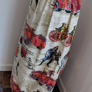 May include: A beige skirt with a repeating pattern of vintage fire engines, firefighters, and dalmatian dogs. The skirt has a high waist and a button closure. The fabric has a slightly textured appearance.