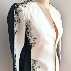 May include: A white and black jacket with beaded embellishments. The jacket has a long sleeve and a collarless neckline. The jacket is on a white mannequin.