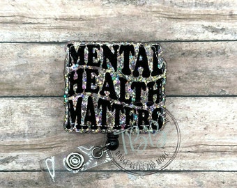 Pode incluir: Um carretel de crachá de glitter prateado com um amuleto quadrado preto que diz "Mental Health Matters".