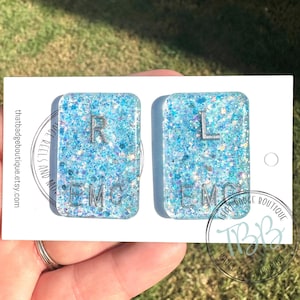 May include: Two rectangular resin badge reels featuring a light blue glitter finish. The left piece displays the letters R and EMC in silver, while the right piece displays the letters L and EMC in silver. Both are mounted on a white display card.