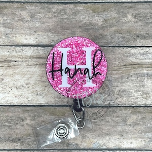 May include: Pink glitter badge reel with a white letter H and the name "Hannah" in black script.