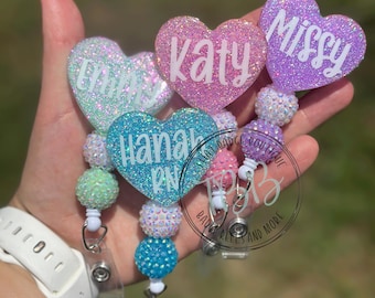 May include: Heart-shaped badge reels in shades of pink, blue, and purple glitter. Each reel features a name in white lettering: Emily, Katy, Missy, and Hannah. The reels have beaded accents and a clear plastic clip.