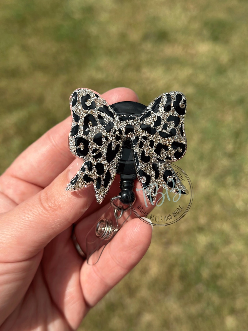 May include: A black and silver leopard print bow badge reel. The bow is made of glittery material and has a black center. The reel has a clear plastic base and a retractable cord.