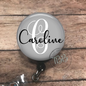 May include: A gray badge reel with the name "Caroline" in a script font, and a large white letter "C". The reel has a clear plastic strap with a decorative gem and a black retractable cord.