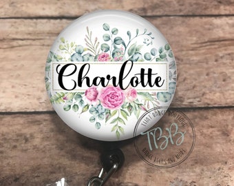 May include: A white round badge reel with a floral design and the name "Charlotte" in a gold frame. The badge reel has a black retractable cord and a clear plastic clip with a crystal.