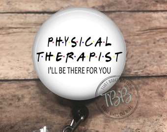 May include: A white badge reel with the words "PHYSICAL THERAPIST" in black, with each letter in a different color. Below the text is "I'LL BE THERE FOR YOU". The reel has a black clip and a clear plastic strap with a decorative gem.