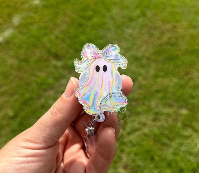 May include: A holographic ghost-shaped badge reel with a bow. The ghost has two black eyes and an iridescent finish. The badge reel is held in a hand against a blurred green background.