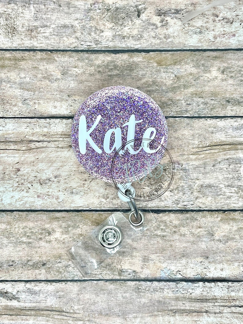 May include: A circular badge reel featuring a purple glitter finish with the name Kate printed in white script lettering. The device includes a retractable cord attached to a clear plastic strap with a metal snap button closure for holding an ID card.