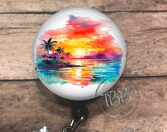 May include: A watercolor sunset scene with palm trees and a reflection of the sun in the water. The image is printed on a round badge reel with a black retractable cord and a clear crystal.