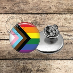 May include: A pinback button with a rainbow flag design. The flag has a black and brown chevron on a white, light blue, and pink background. The rainbow colors are red, orange, yellow, green, blue, indigo, and violet.