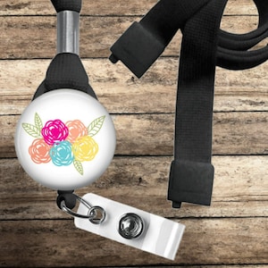 May include: A white retractable badge reel featuring a floral design in pink, orange, blue, and yellow. It's attached to a black lanyard and a white plastic badge holder. The background is a wooden surface.
