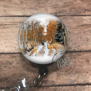 May include: A white round badge reel with a picture of a snowy forest scene with two deer. The text "That Badge Boutique" and "Badge Reels and More" is printed on the reel.