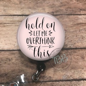 May include: Pink round badge reel with a black and white design that says "hold on let me overthink this". The badge reel has a clear plastic badge holder and a silver swivel clip.