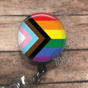May include: A round badge reel with a black retractable cord and a clear crystal charm. The badge holder features a colorful design with a rainbow flag and a black chevron. The text "That Badge Boutique" and "Badge Reels and More" is printed in a circle around the design.