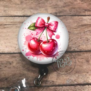 May include: A white round badge reel with a pink cherry design. The cherries have a pink bow on top and are surrounded by pink watercolor splatters. The badge reel has a clear plastic badge holder and a silver swivel clip.