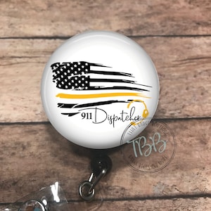 May include: A white badge reel with a black and white American flag with a yellow stripe across the middle. The text "911 Dispatcher" is printed on the badge reel. The badge reel is attached to a black retractable cord.