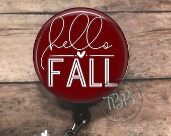 Peut inclure: Un porte-badge rond rouge avec un motif blanc qui dit "hello fall" avec un petit cœur au milieu. Le porte-badge est sur un fond en bois.