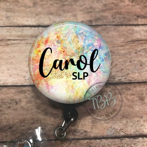 May include: A colorful, abstract badge reel with a gold glitter background. The name "Carol SLP" is written in black text on the front of the reel.