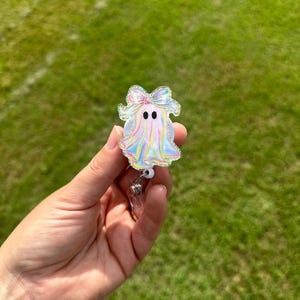 May include: A holographic ghost-shaped badge reel with a bow on top. The badge reel has two black dot eyes and a silver retractable clip. The ghost is held in front of a green grassy background.