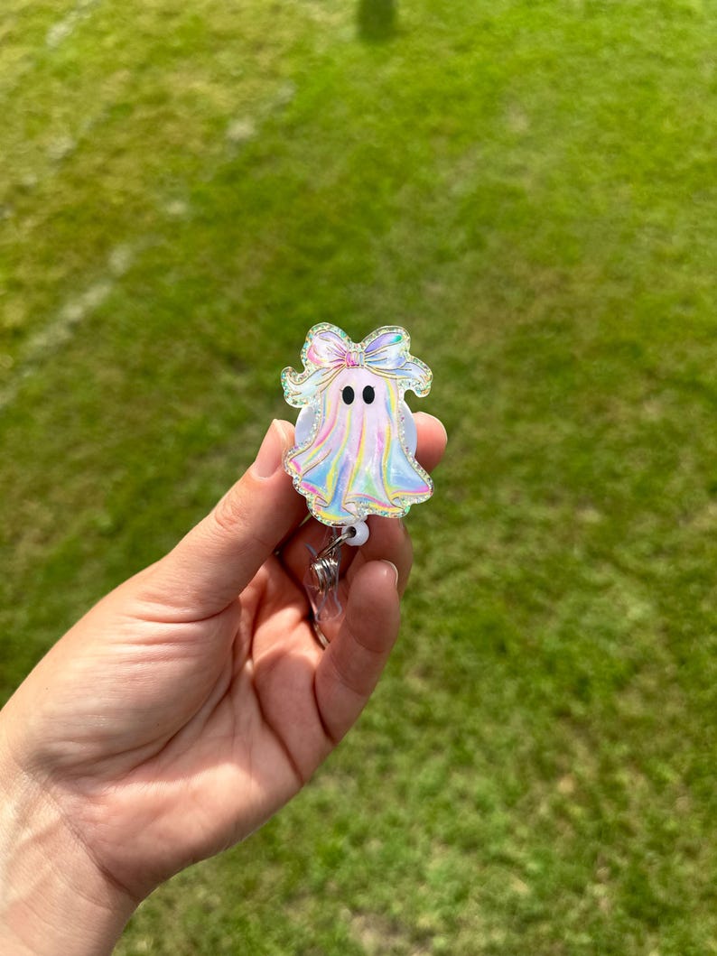 May include: A holographic ghost-shaped badge reel with a bow on top. The badge reel has two black dot eyes and a silver retractable clip. The ghost is held in front of a green grassy background.