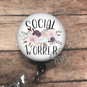 May include: A white badge reel with a floral design and the text "SOCIAL WORKER". The badge reel is attached to a black retractable cord and a clear plastic badge holder with a crystal.