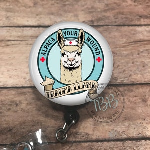 May include: A white round badge reel with a blue and white circle design. The circle has the text "Alpaca Your Wound" with a red cross on each side. Inside the circle is a cartoon image of a llama wearing a nurse's hat. Below the circle is a banner with the text "Trauma Llama".