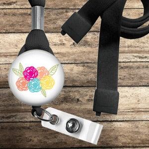 May include: A white retractable badge reel with a floral design in pink, orange, blue, and yellow. The reel is attached to a black lanyard and a white plastic badge holder. The background is a wooden surface.