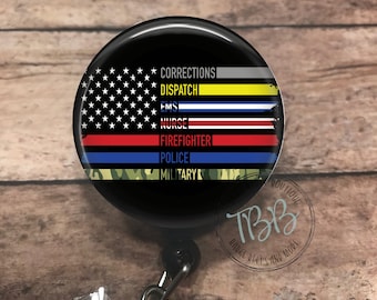 Op de afbeelding: Een zwarte intrekbare badgehouder met een ronde knop met een Amerikaanse vlag met dunne rode, witte en blauwe strepen en de woorden "Corrections, Dispatch, EMS, Nurse, Firefighter, Police, Military" erop gedrukt.